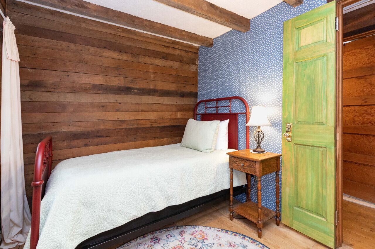 Une petite chambre à coucher accueillante, avec un lit simple, une table de chevet, une lampe et une porte verte.
