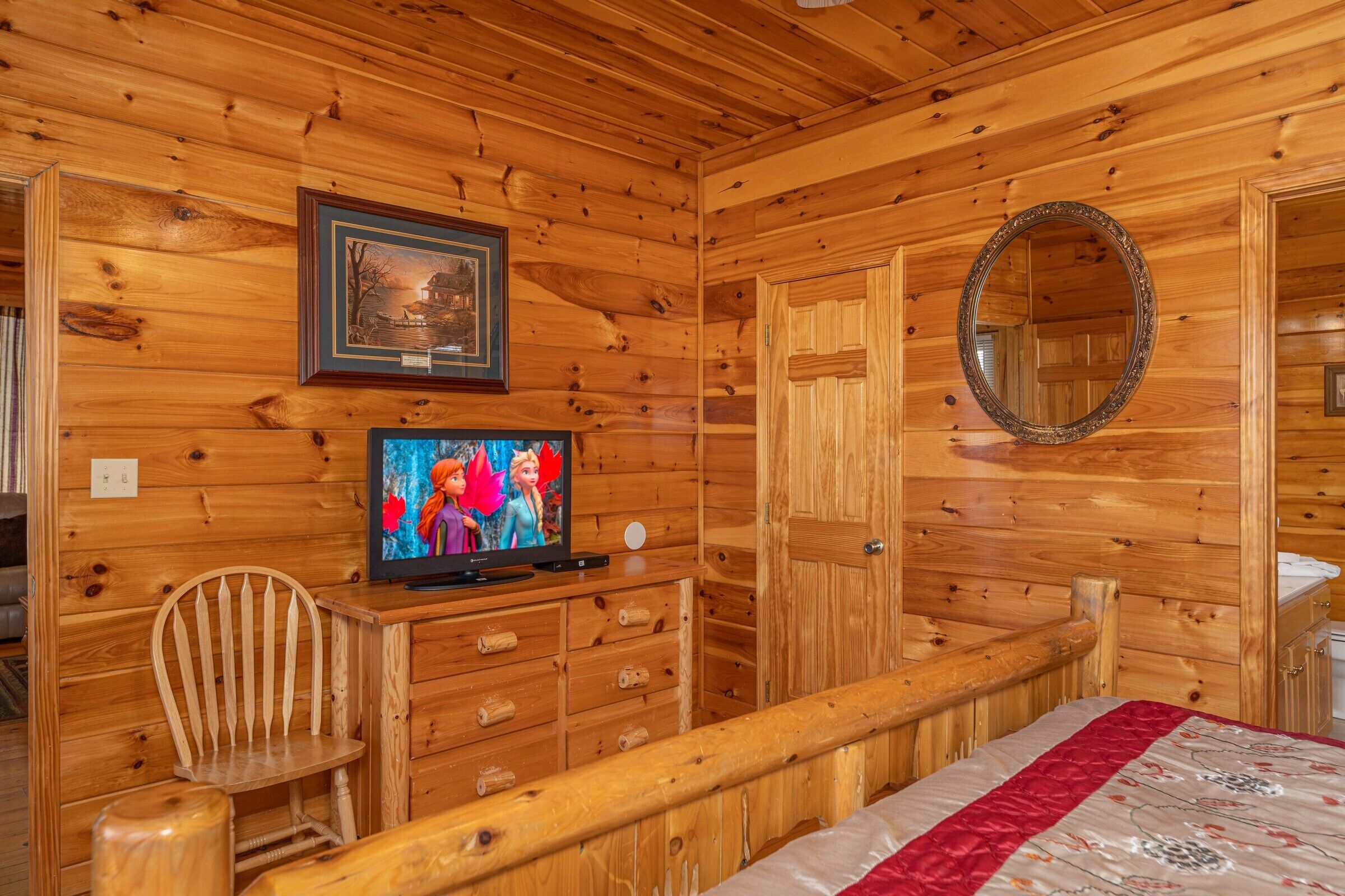 Een houten kamer met een televisie, een houten stoel, een bed met een rode en witte dekbedovertrek en een spiegel aan de muur.