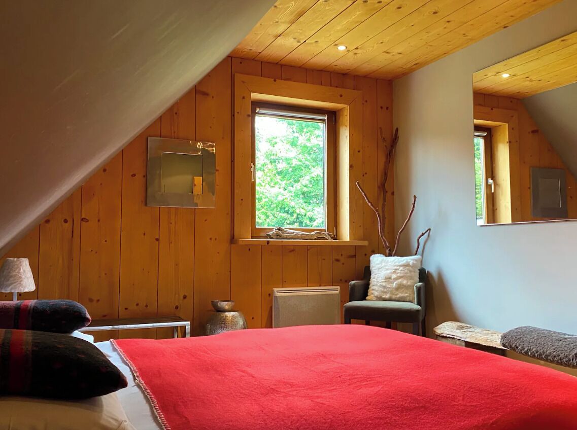 A cozy room with a wooden ceiling, a large window, a bed with a red blanket, a chair, and a small table.
