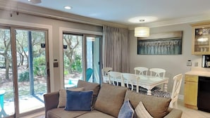 Ein Wohnzimmer mit einer Couch, einem Esstisch mit Stühlen, einer Küchenzeile und großen Schiebeglastüren, die zu einem Garten führen.
