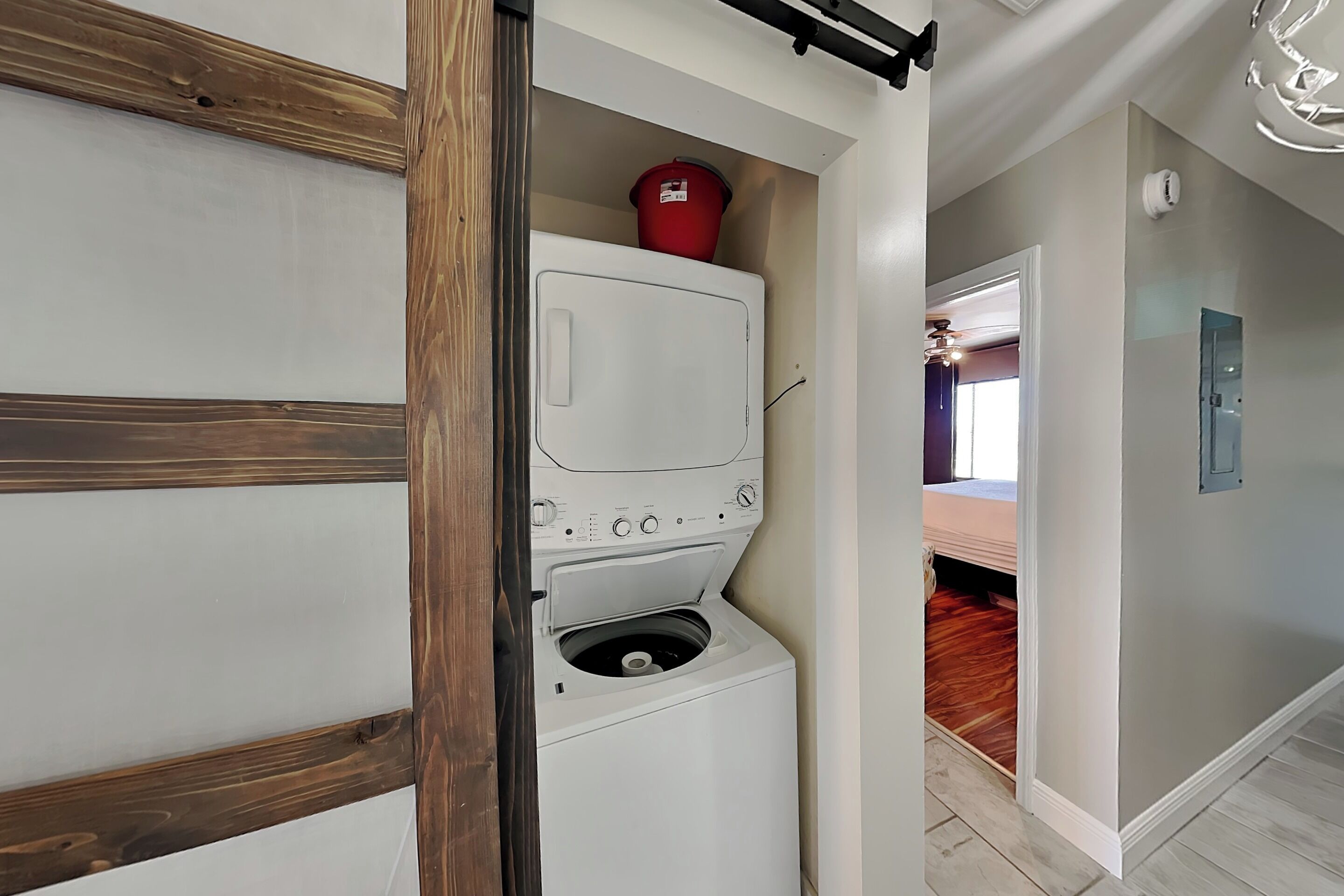 A laundry room with a washer and dryer, a red bucket on the dryer, and a hallway leading to a bedroom.