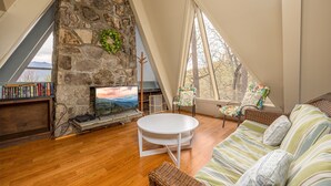 A cozy loft bedroom with a stone fireplace, a round white table, a sofa with striped cushions, and large windows offering a view of the outdoors. - Your Family Reunion Getaway (GATLINBURG)