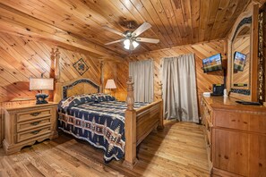 Una stanza con rivestimento in legno, un letto, una cassettiera, un comodino, una ventola a soffitto e una televisione.