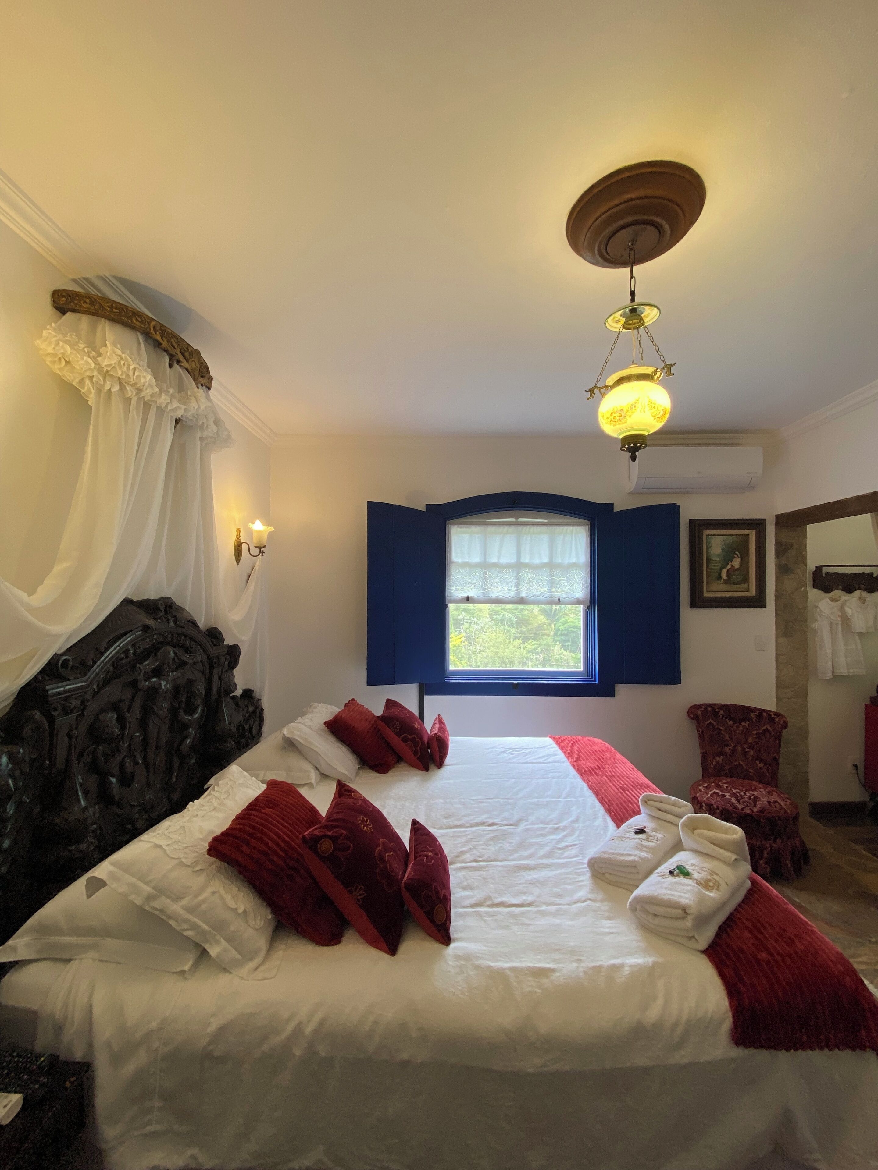 Una habitación de hotel con una cama grande, una ventana con contraventanas azules, un candelabro y una silla roja.