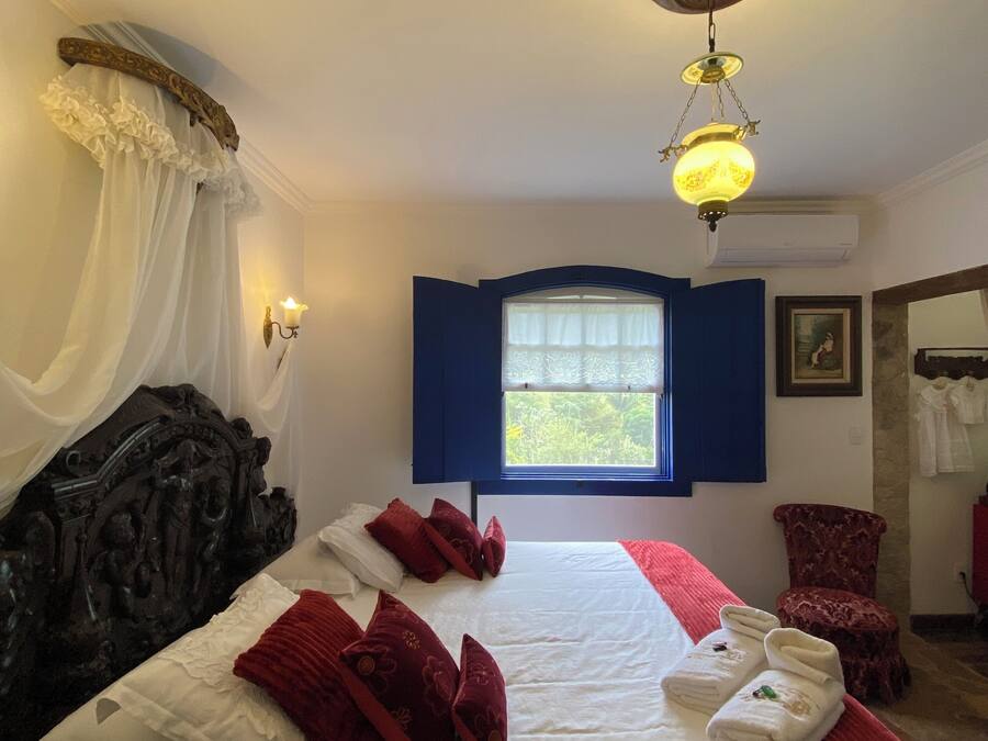 Una habitación de hotel con una cama grande, una ventana con contraventanas azules, un candelabro y una silla roja.