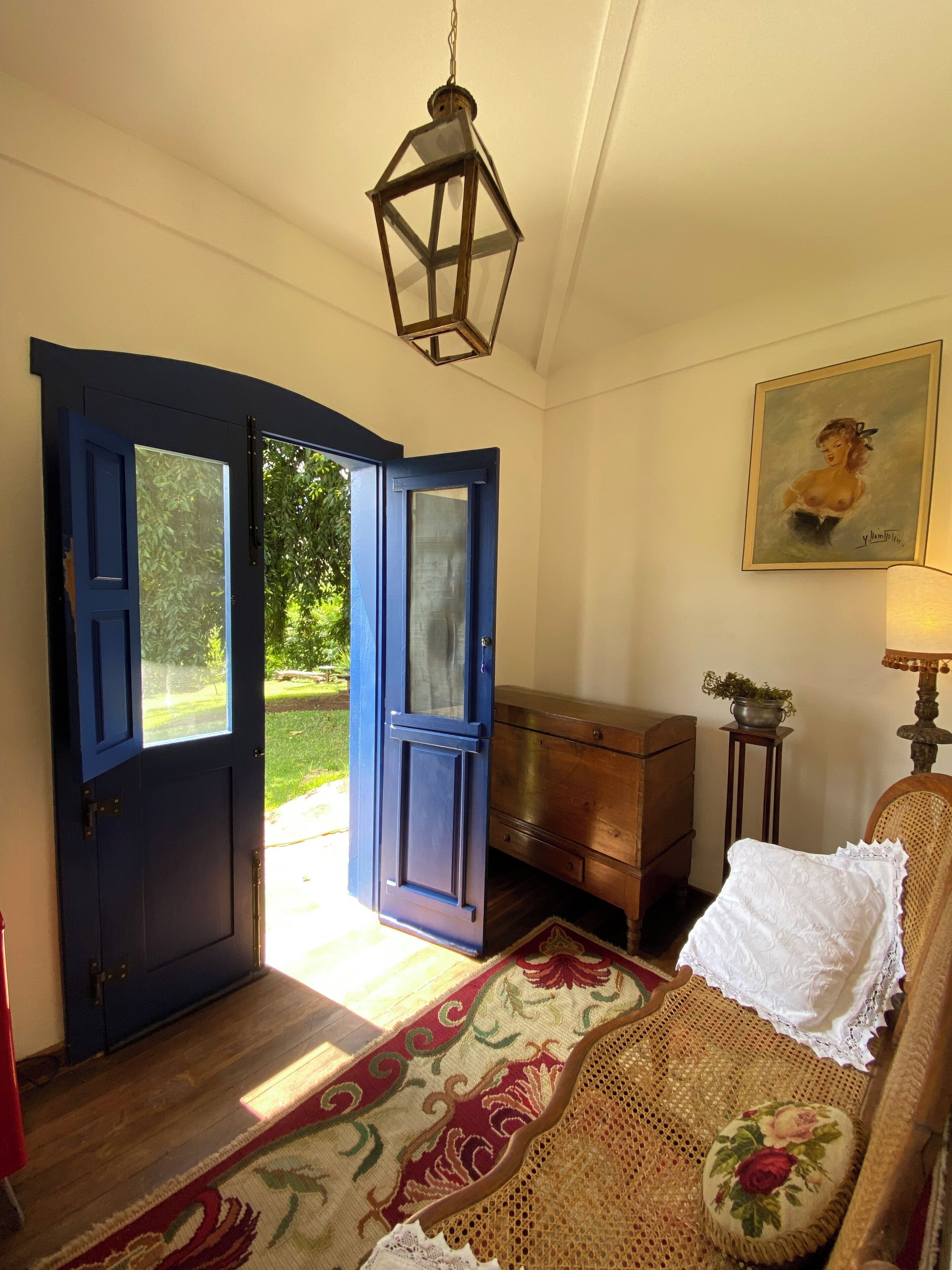 A room with a large blue door, a wooden dresser, a bed with a floral bedspread, a hanging lantern, and a painting on the wall.