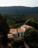 Una casa con techo de tejas, rodeada de abundante vegetación y colinas al fondo.