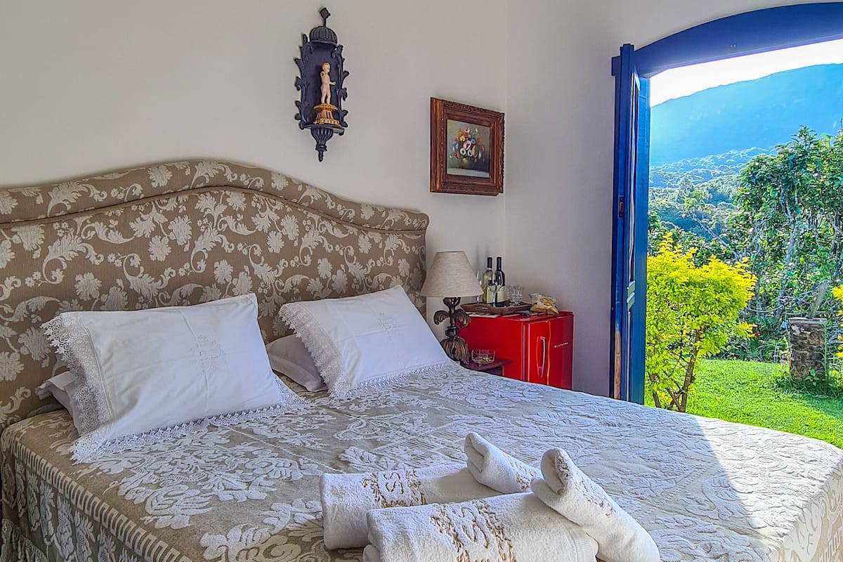 A neatly made bed with a patterned bedspread and pillows, a bedside table with a lamp and a small decorative item, and a view of a lush garden through an open door.