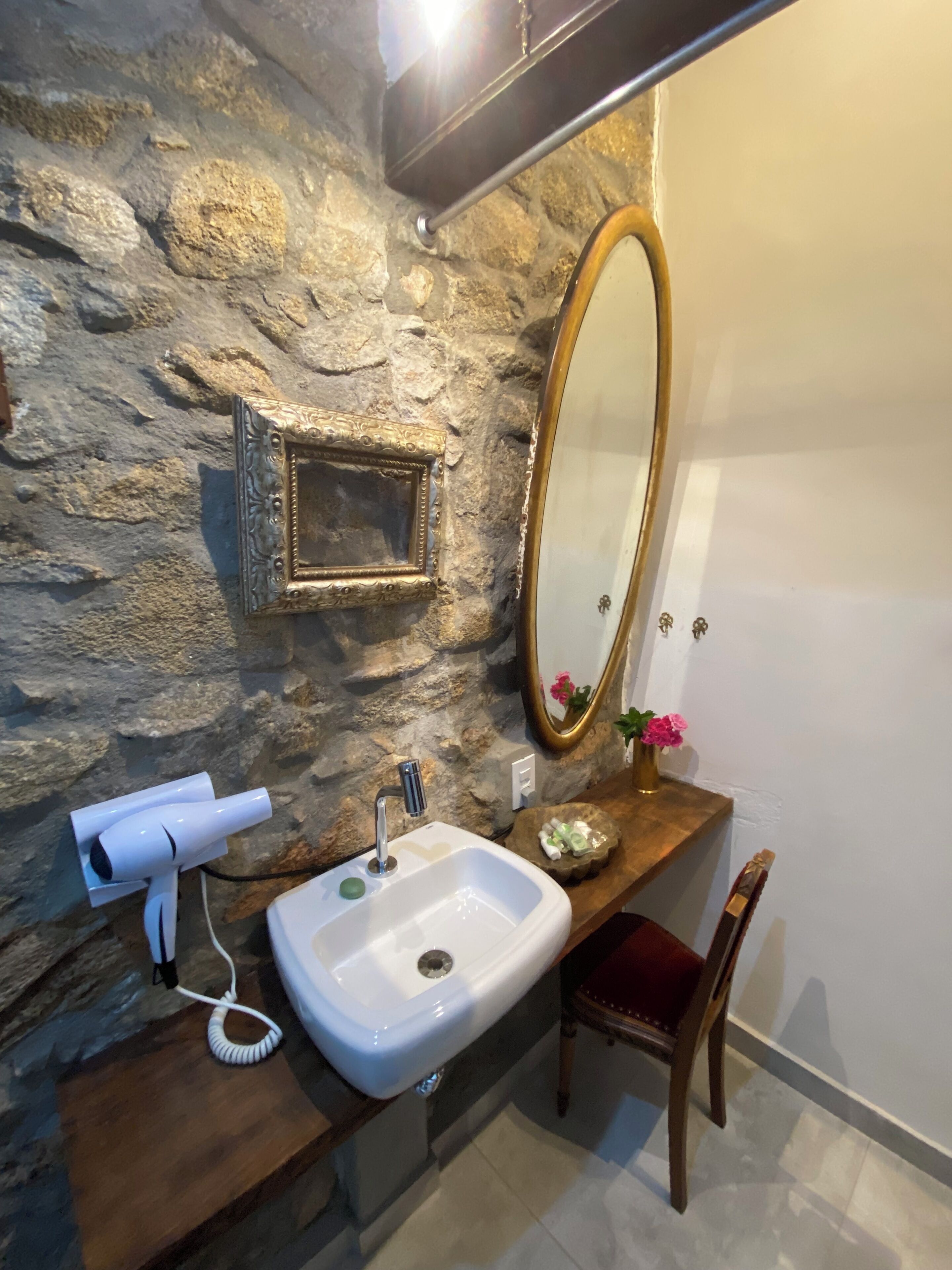 A bathroom with a stone wall, a white sink, a hairdryer, a mirror, and a wooden chair.