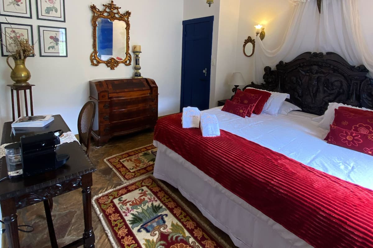 A bedroom with a large bed, a dresser, a desk, and a chair. There are framed pictures on the wall and a mirror above the dresser.