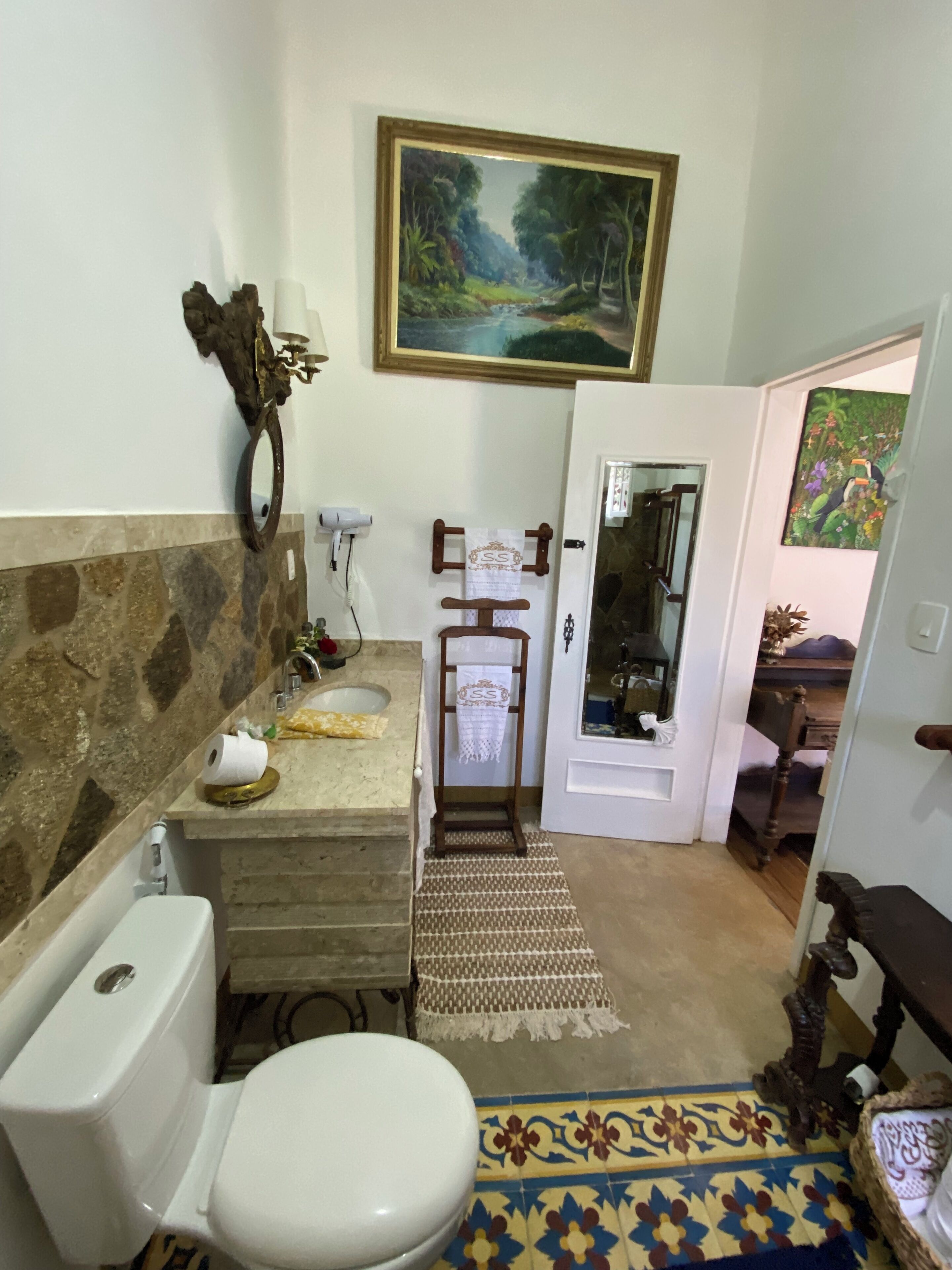 A bathroom with a stone wall, a sink, a toilet, a rug, and a painting above the mirror.