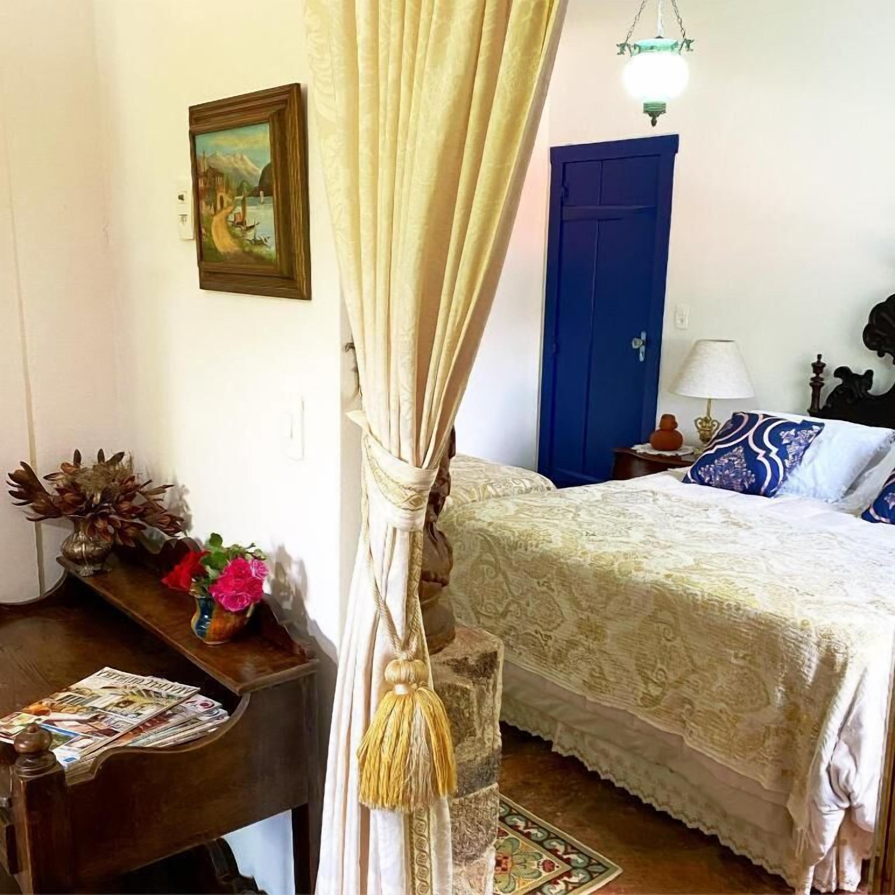 Un dormitorio con cama, un escritorio con lámpara, un cuadro en la pared y una cortina.