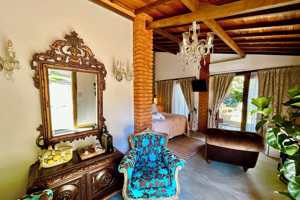 A room with a large ornate mirror, a vintage armchair, a bed with a pink bedspread, and a chandelier.