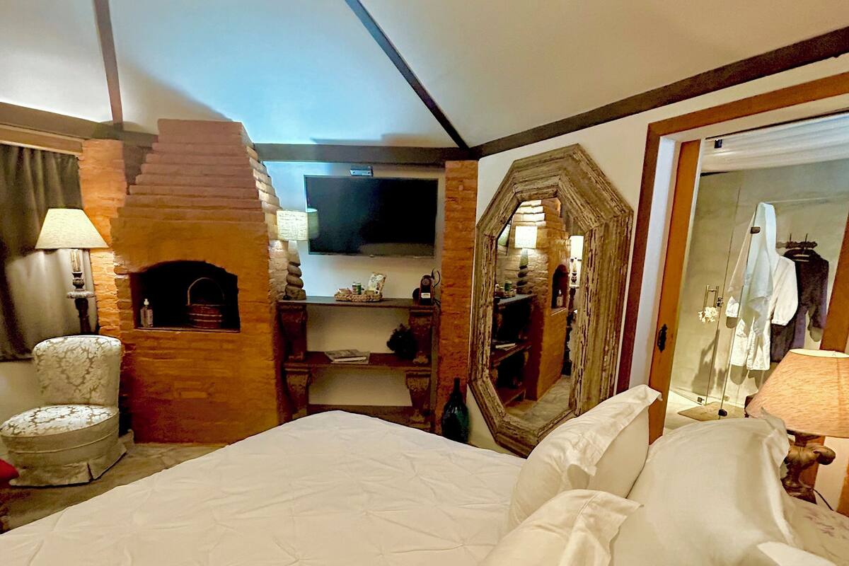 A hotel room with a large bed, a chair, a fireplace, a mirror, and a television.