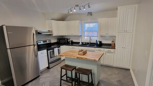 A modern kitchen with white cabinets, stainless steel appliances, a wooden island with stools, and a window providing natural light. - Spacious, Fully-Stocked, Walking distance to Downtown - With Comfy Mattresses! (Bardstown)