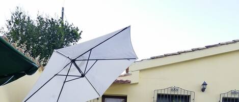 A patio with stone flooring, a table with chairs, a white umbrella, and a small garden area with a bush.
