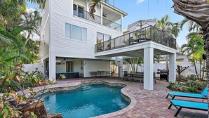 Una casa de dos pisos con piscina, mobiliario exterior y un patio cubierto.