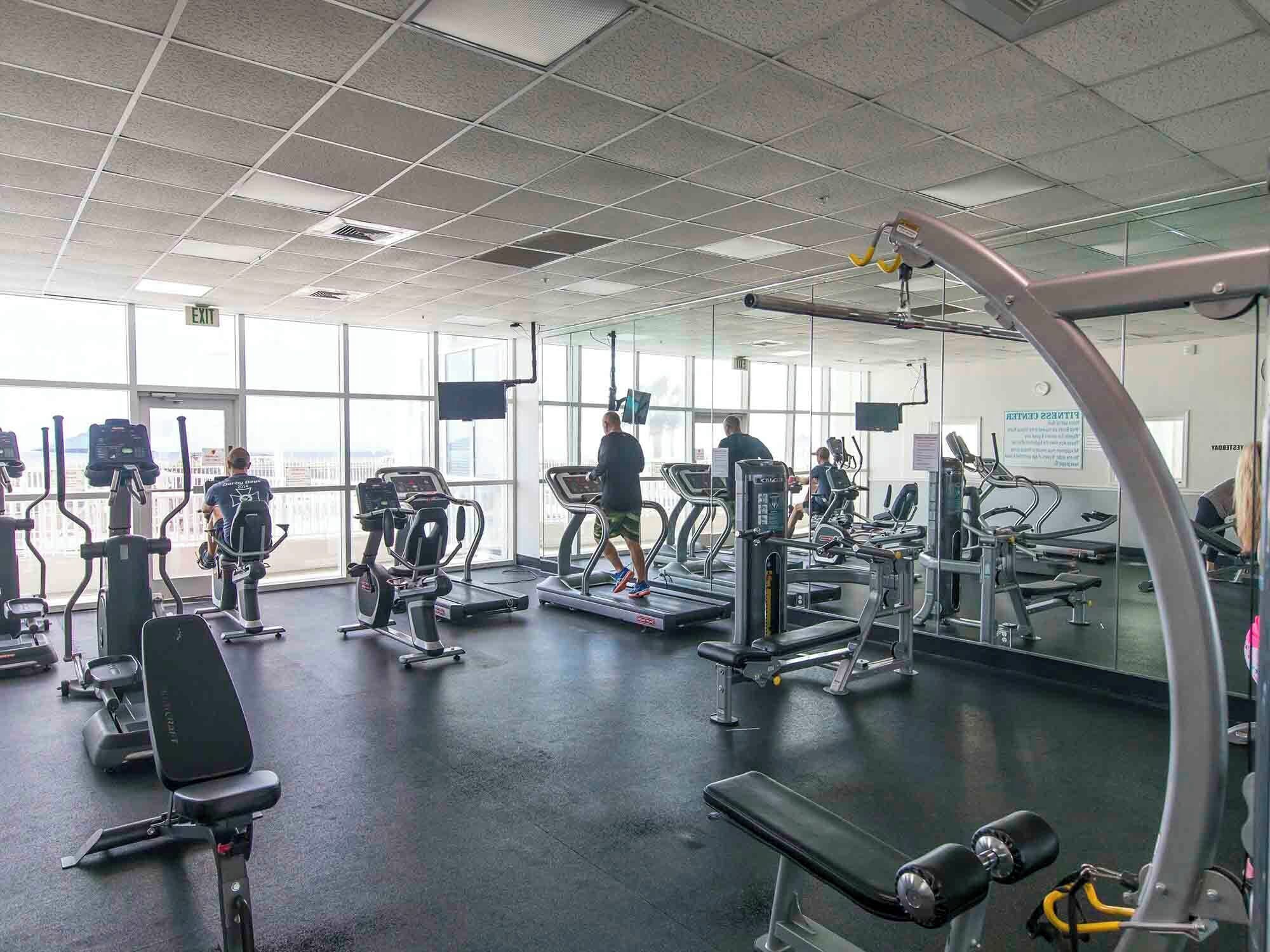 A gym with various exercise machines, large windows, and a mirrored wall.