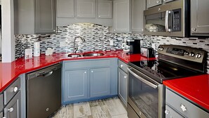 A modern kitchen with grey cabinets, a red countertop, a stainless steel sink, a microwave, and an oven. - Paradise Isle #15A by Southern Vacation Rentals (Gulf Shores)