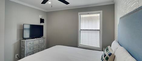 Un dormitorio con cama, ventilador de techo, televisor sobre una cómoda y ventana con persianas.