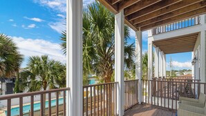 Un espace extérieur abrité doté d’une terrasse en bois, de colonnes blanches et offrant une vue sur une piscine et des palmiers.