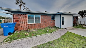A single-story brick house with a small attached garage, a blue trash bin, and a paved walkway leading to the entrance. - Seashell Cottage by Resort Collection (Panama City Beach)