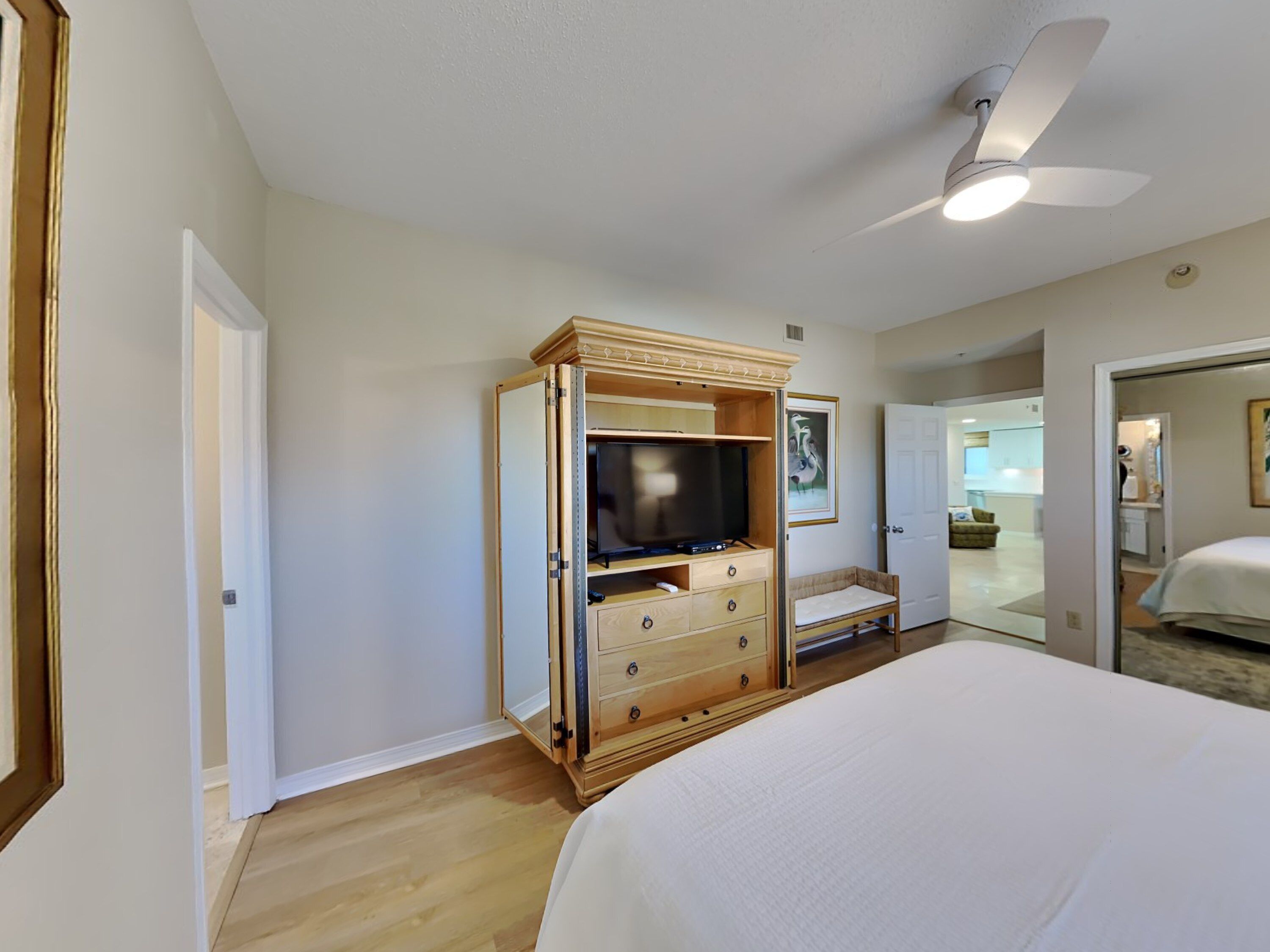 A bedroom with a bed, a wooden TV stand, a ceiling fan, and a door leading to a bathroom.