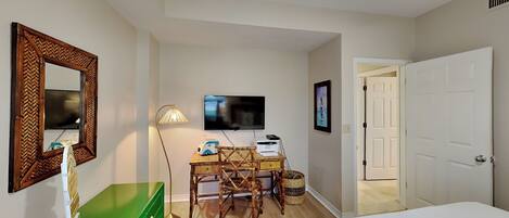 A hotel room with a bed, a green dresser, a desk with a lamp, a television, and a door.
