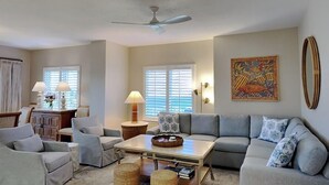 A spacious living room with a gray sofa, a coffee table, and a ceiling fan. There are two windows with blinds, a decorative wall piece, and a lamp on a side table. - South Harbour #2F by Southern Vacation Rentals (Pensacola Beach)