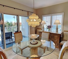 A dining area with a glass table, chairs, a chandelier, and a view of the outdoors.