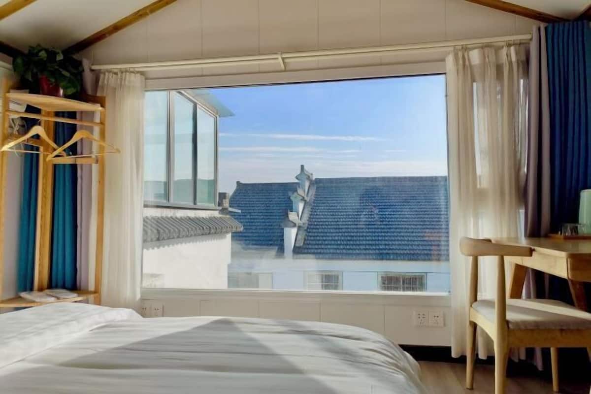 Une chambre avec une grande fenêtre, un lit, une chaise en bois et un portant sur lequel sont suspendus des vêtements.