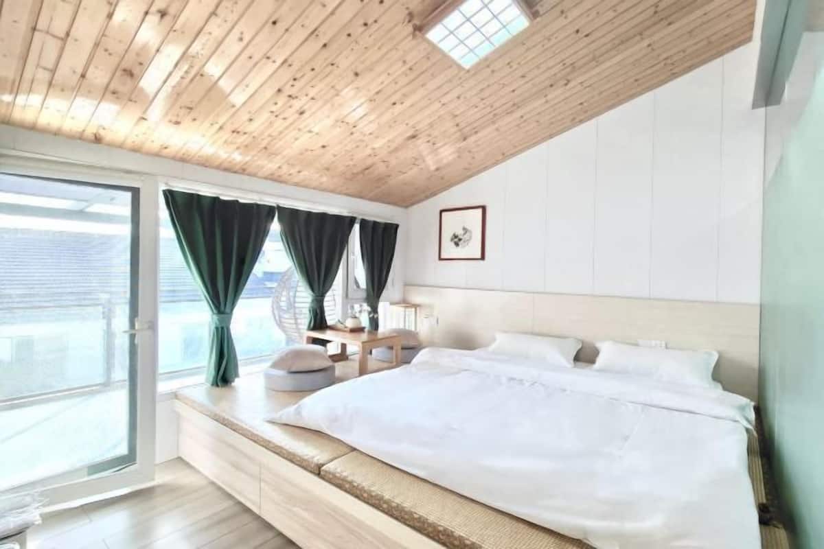 Une chambre avec un plafond en bois, un grand lit, une fenêtre avec des rideaux et un tableau au mur.