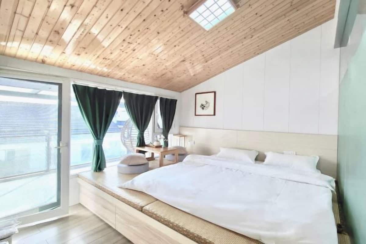 Une chambre avec un plafond en bois, un grand lit, une table de chevet et une fenêtre avec des rideaux.
