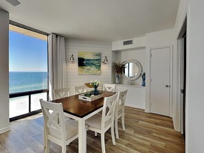 A dining area with a wooden table and white chairs, a painting on the wall, and a view of the ocean.