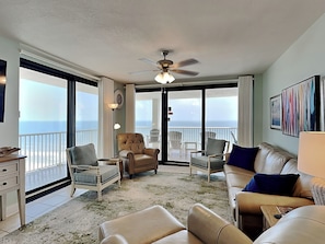 A spacious living room with a large window overlooking the ocean, a ceiling fan, and a variety of seating options including a sofa, chairs, and a small table.