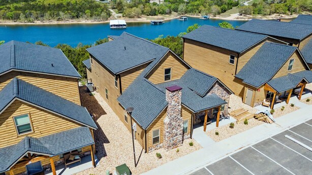 A cluster of log cabins with a stone chimney, a parking lot, and a lake with boats. - Lakefront Cabin–Pool, Private Hot, Tub & Fireplace (Branson)