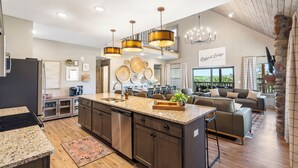 A spacious kitchen with granite countertops, stainless steel appliances, and a large island. There is a dining area with a gray sofa and a stone fireplace. - Lakefront Cabin–Pool, Private Hot, Tub & Fireplace (Branson)