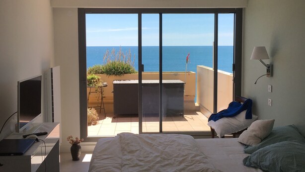 A bedroom with a bed, a desk with a computer, a TV, and a view of the sea through large sliding doors. - Terrasses sur la mer (Sète)