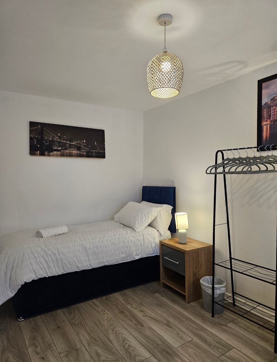 A bedroom with a bed, a nightstand, a hanging light fixture, a wall-mounted picture, and a metal rack.