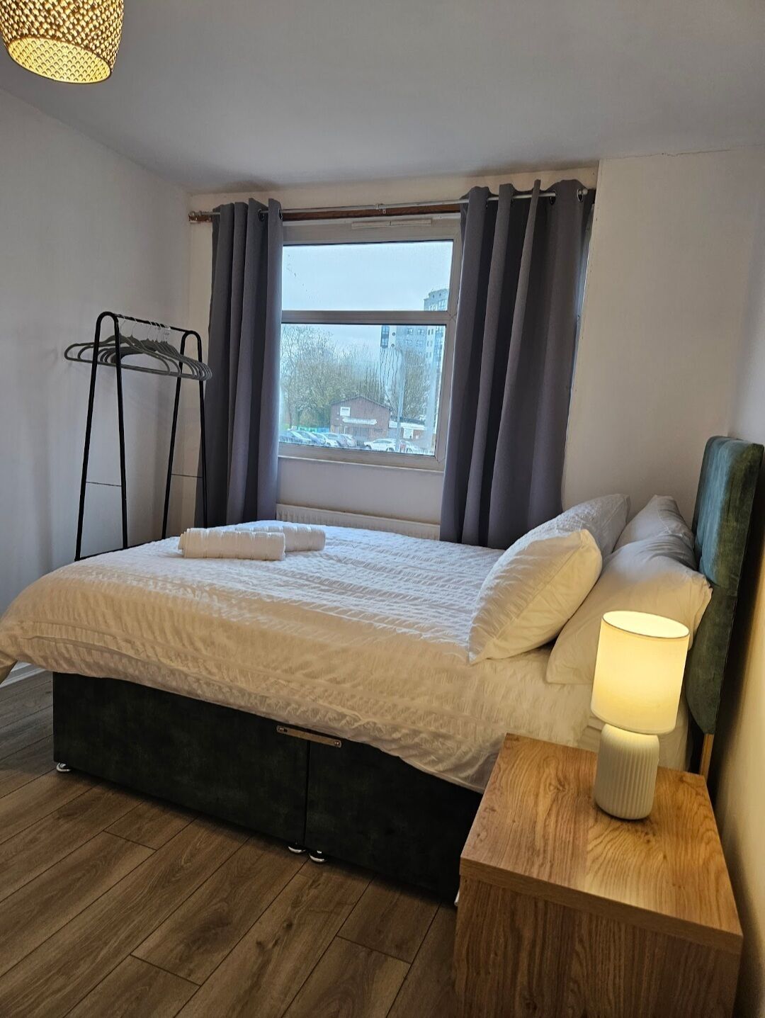 Un dormitorio con cama, mesita de noche con lámpara, ventana con cortinas y perchero.