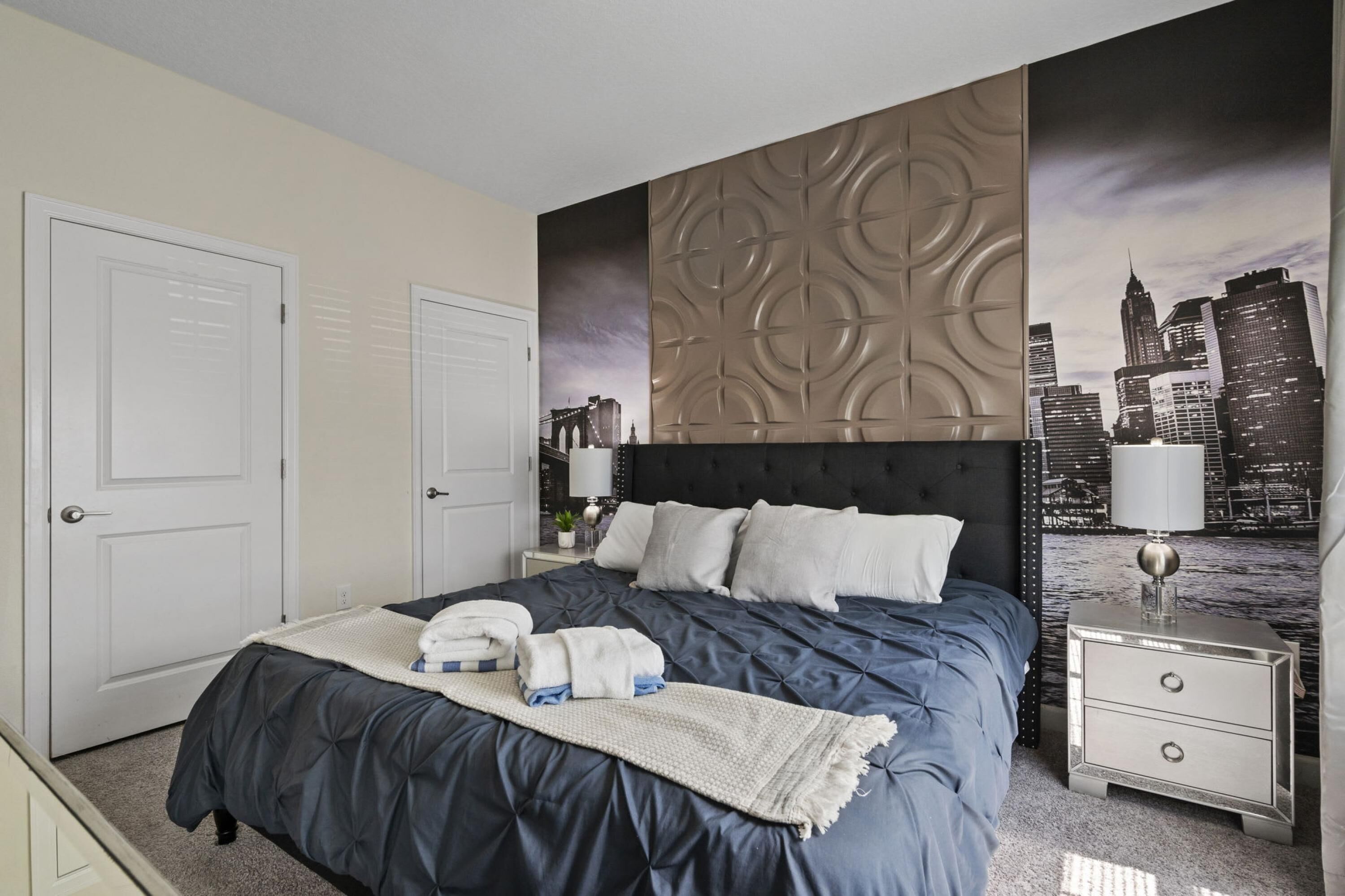 A hotel room with a large bed, a nightstand with a lamp, and a cityscape mural on the wall.