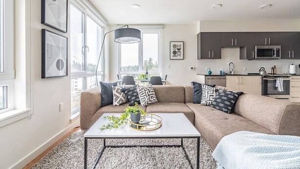 A modern living room with a sectional sofa, a coffee table, and a view of the city. - 2BR | Gym, Views | Bellevue | Sophari Parkside F (Bellevue)