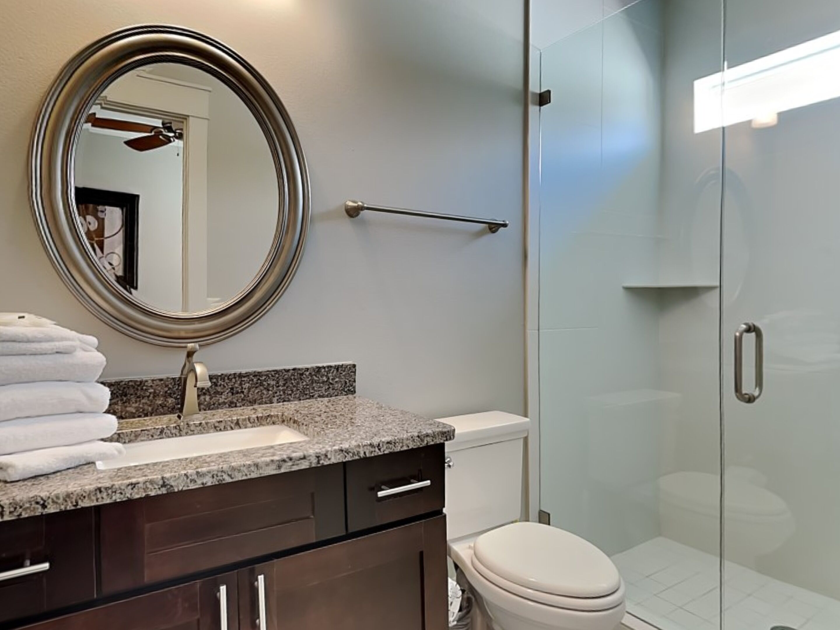 A modern bathroom with a granite countertop, a round mirror, a towel rack, and a glass shower enclosure.