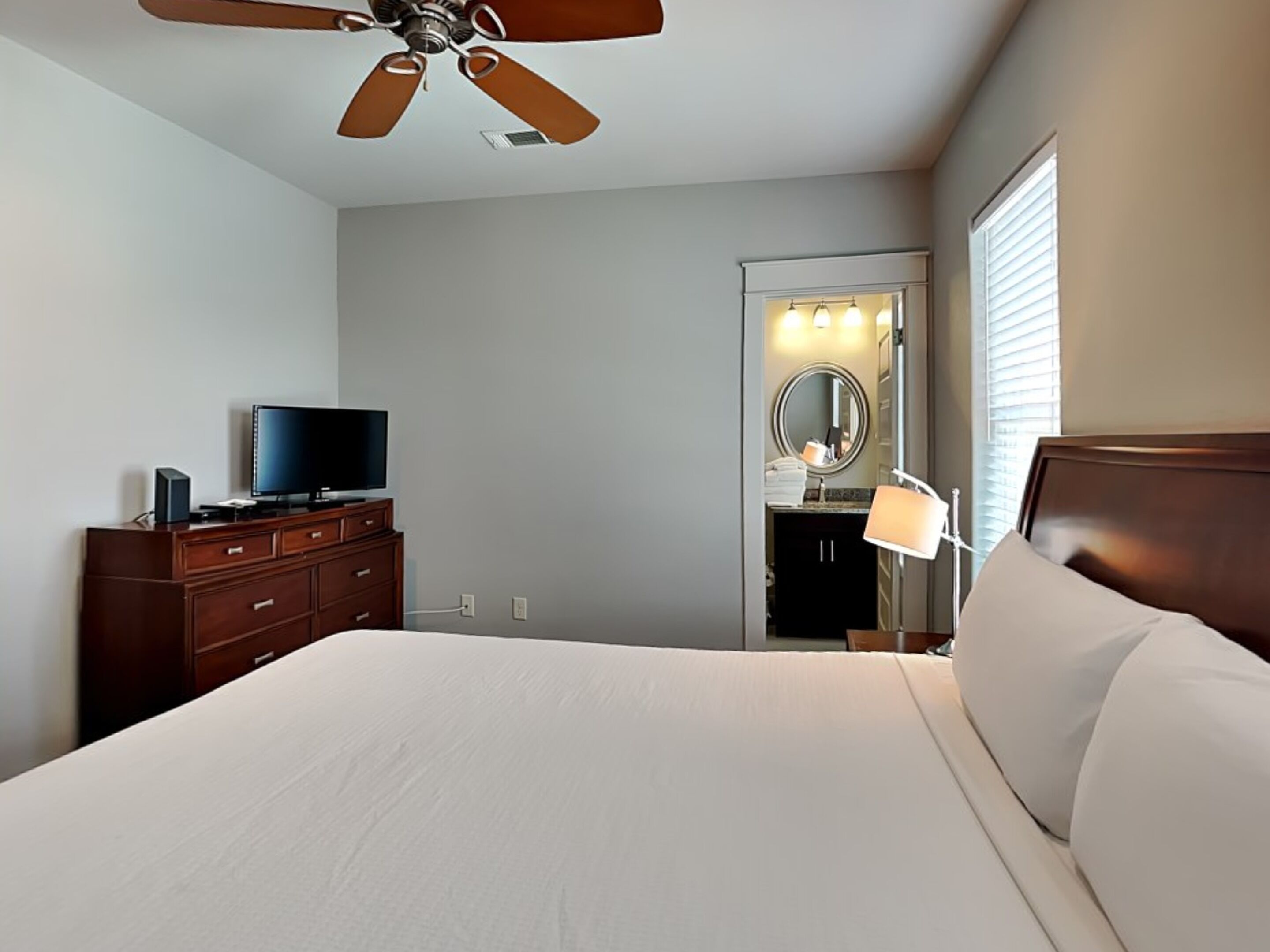A hotel room with a bed, a TV on a wooden dresser, a ceiling fan, a bathroom with a mirror and sink, and a window with blinds.