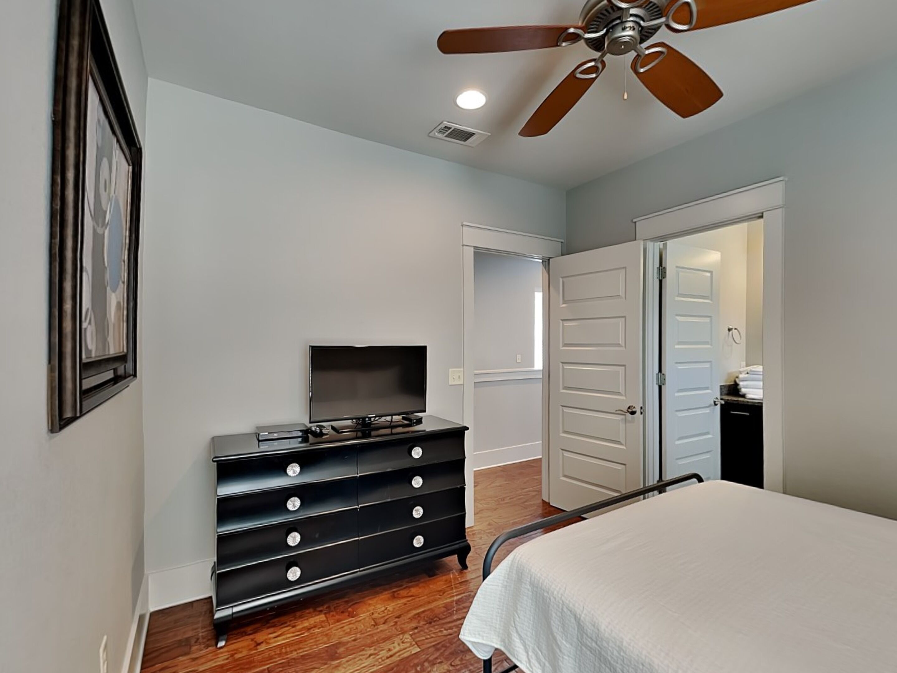A bedroom with a bed, a dresser, a television, a ceiling fan, and a door leading to another room.