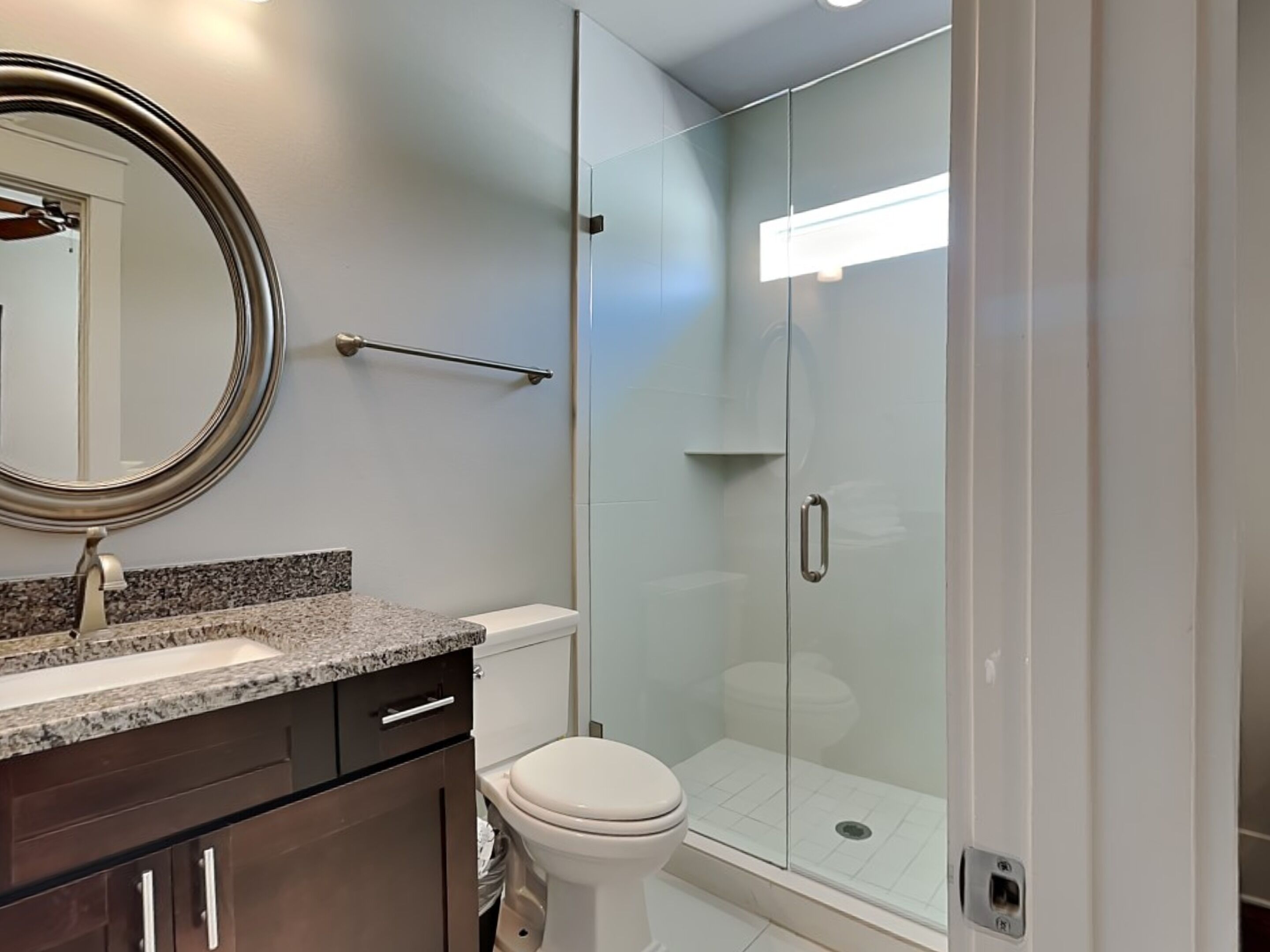 A modern bathroom with a granite countertop, a toilet, a glass shower enclosure, and a mirror.