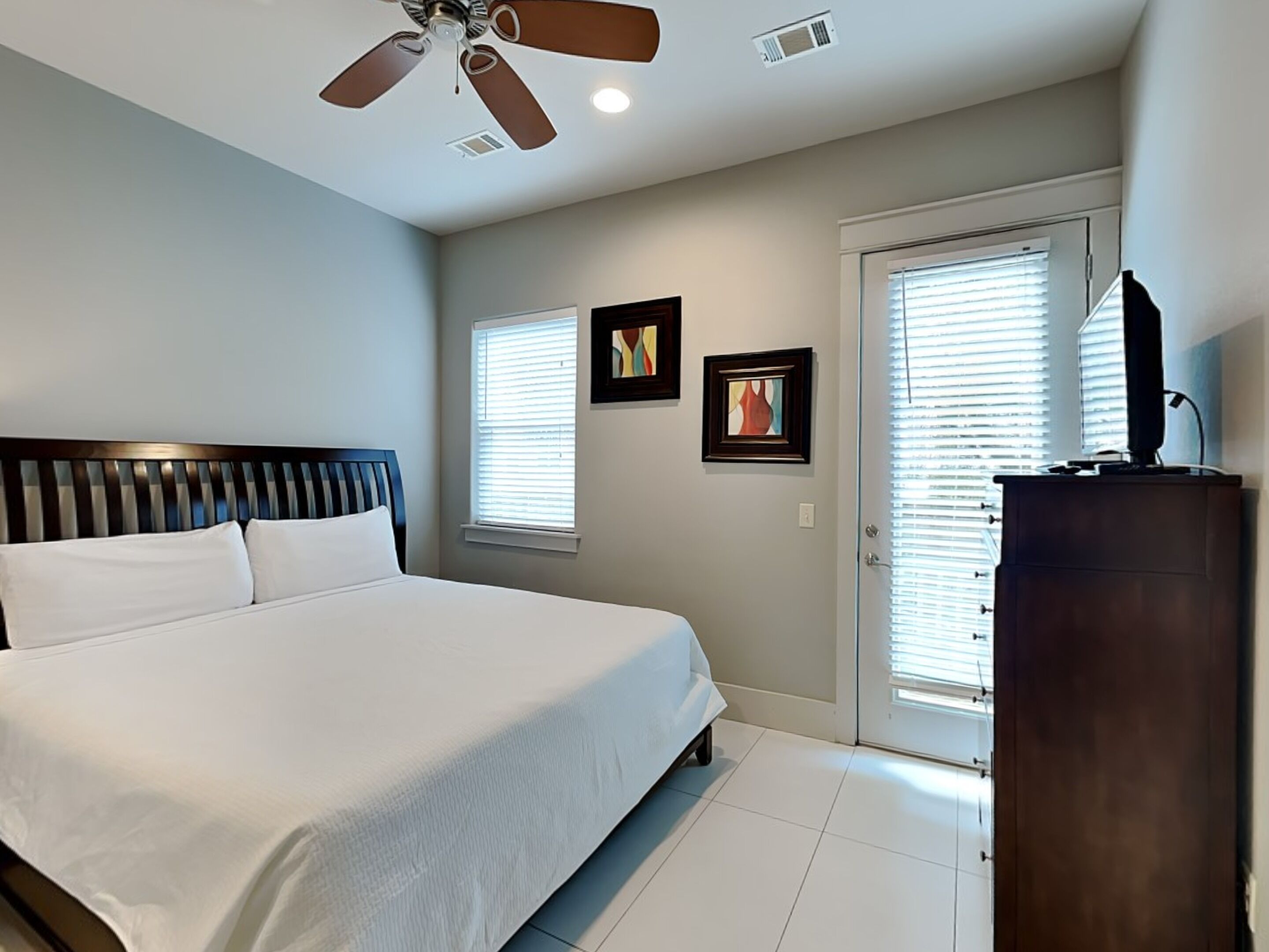 A bedroom with a bed, ceiling fan, two windows with blinds, and a dresser.