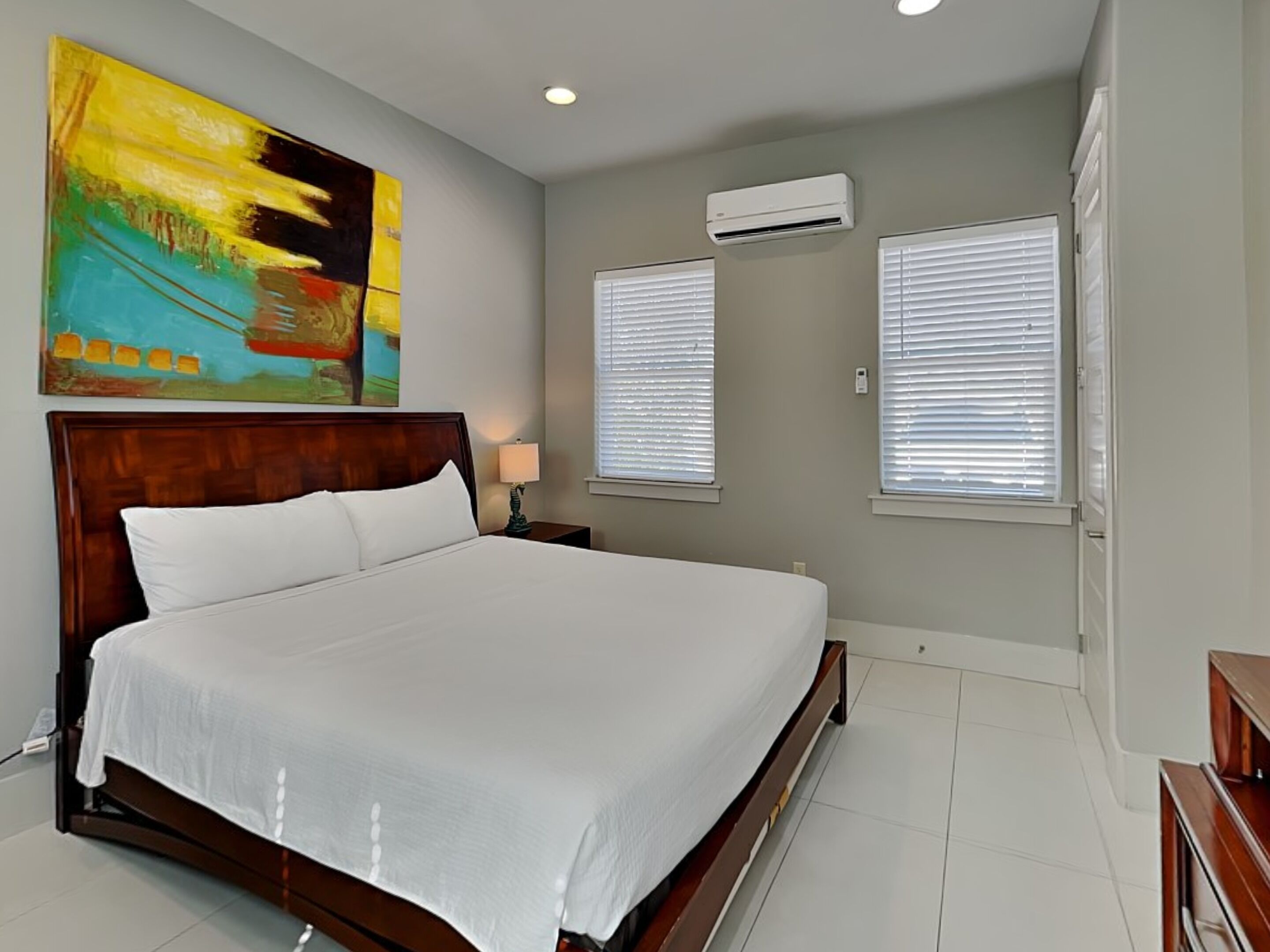 Une chambre d’hôtel avec un grand lit, un appareil de climatisation, deux fenêtres équipées de stores et un tableau abstrait aux couleurs vives accroché au mur.