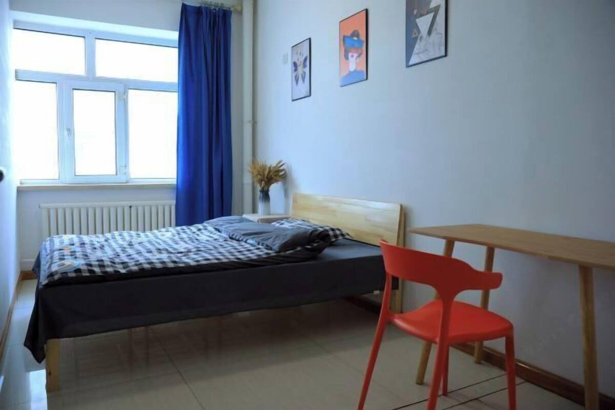 Ein Schlafzimmer mit einem Bett, einem roten Sessel, einem Holztisch und einem Fenster mit blauen Vorhängen.