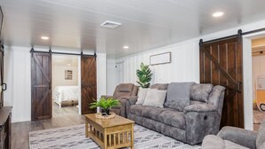 Een woonkamer met een grijze bank, een houten salontafel en een kamerkleed met een patroon. Er is een schuifdeur in boerderijstijl en een lijst met een foto aan de muur.